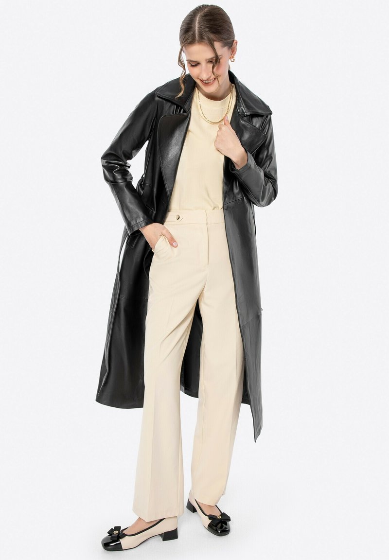 Cappotto lungo in pelle nera, top beige, pantaloni su misura chiari e scarpe con tacco a blocco nere e beige con dettaglio a fiocco.