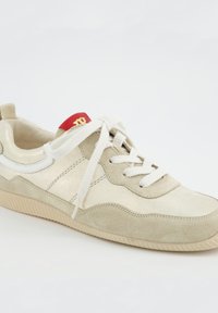Beige semišové a kožené tenisky s bílými tkaničkami, světle béžovou podrážkou a červeným štítkem na jazyku se zlatým logem na bílém pozadí.