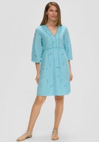 Helles blaues Kleid mit 3/4-Ärmeln, V-Ausschnitt und bestickten Mustern. Das Kleid hat eine geraffte Taille und einen lässigen Schnitt. Es wird mit flachen Sandalen getragen.