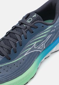 Zapatilla deportiva gris con parte superior de malla y sintética, acentos verdes y suela acolchada. Presenta cordones prominentes y un logo en la lengüeta.