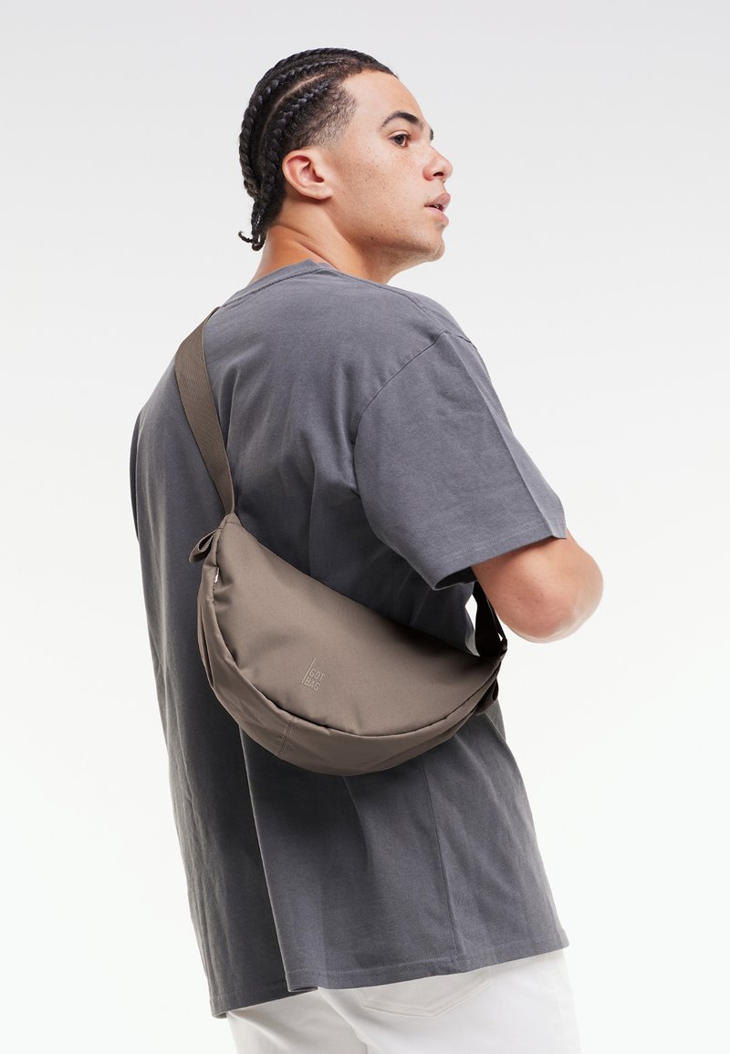 Borsa a tracolla marrone realizzata in tessuto morbido, con una forma curva e una singola tracolla regolabile, indossata sopra una camicia grigia su uno sfondo semplice.