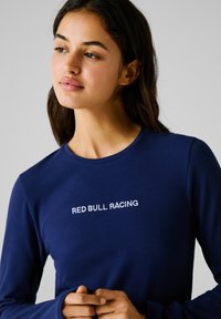 Mlada ženska s temnimi lasmi nosi mornarsko modro majico z dolgimi rokavi, na prsih pa ima belo natisnjen napis "RED BULL RACING", rahlo pogleda vstran.