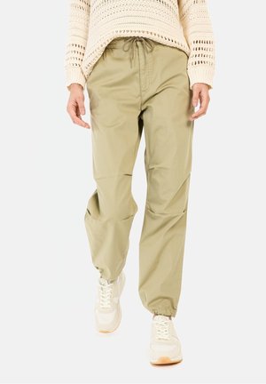Person trägt beige Hose mit Kordelzug und elastischen Bündchen, weiße Sneaker und einen cremefarbenen Strickpullover, geht vorwärts auf einem einfarbigen Hintergrund.