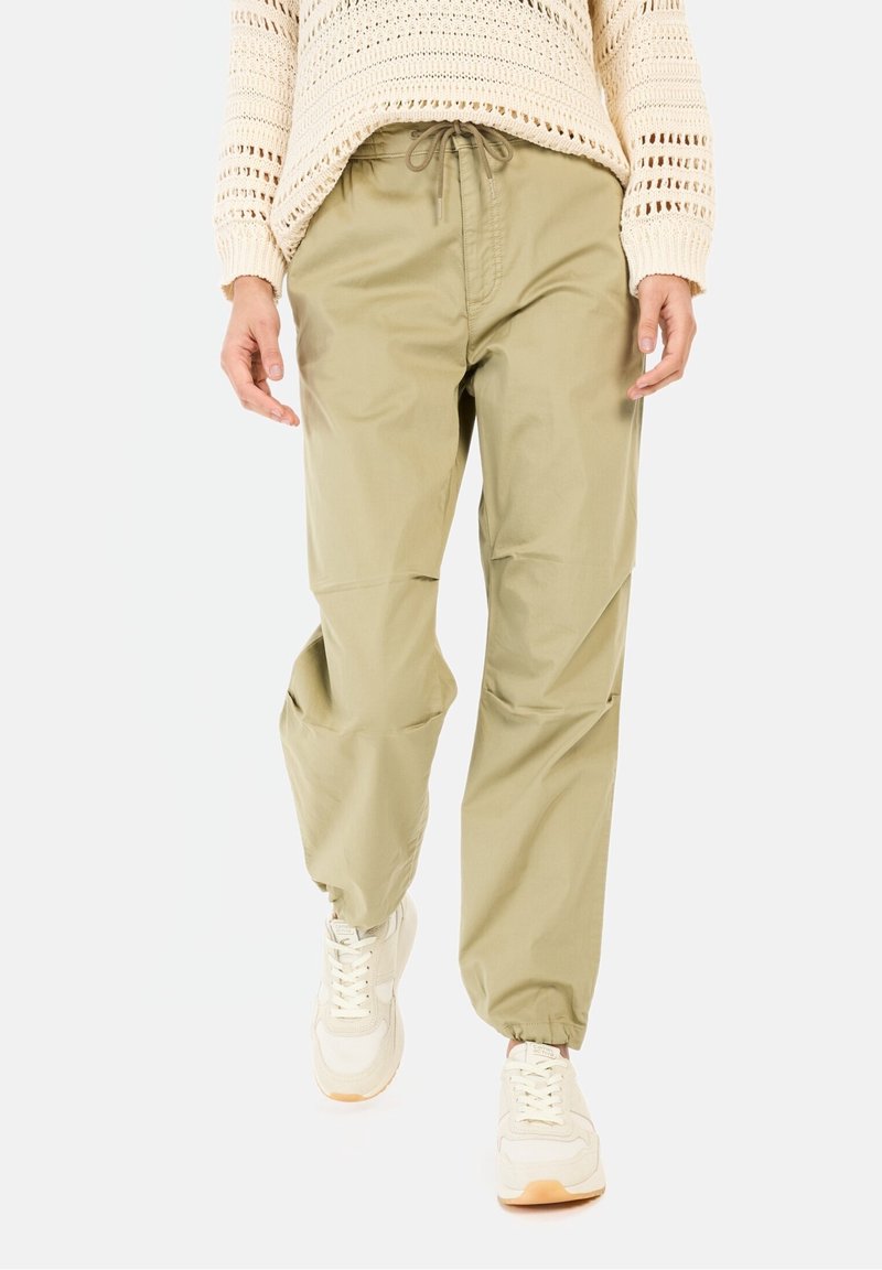 Personne portant un pantalon beige à cordon avec des poignets élastiques, des baskets blanches et un pull en maille crème, marchant vers l'avant sur un fond uni.