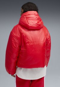 Rode pufferjack met een hoge kraag en capuchon, met een gladde, glanzende textuur. Het ontwerp is cropped met brede mouwen, wat de warmte benadrukt.