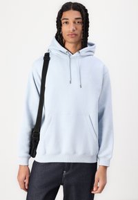 Hellblaues Hoodie aus weichem Material mit einer vorderen Kängurutasche, Kapuze mit Kordelzug und gerippten Bündchen. Kombiniert mit dunklen Jeans.