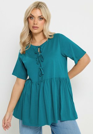 Mujer rubia con blusa peplum de manga corta en color verde azulado con detalles de lazo en el frente y vaqueros azul claro, posando frente a un fondo liso.