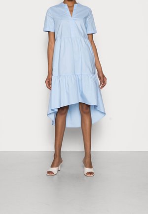 Nainen, jolla on päällään vaaleansininen lyhythihainen mekko, jossa on korkeampi takahelma kuin edessä, ja valkoiset avojalkaiset korkokengät, seisomassa harmaalla lattialla.