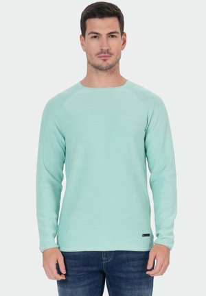 Homme portant une chemise à manches longues vert clair et un jean bleu, debout devant un fond blanc uni.
