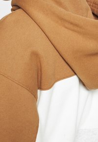 Felpa con cappuccio marrone e bianca caratterizzata da una consistenza morbida, con un design a blocchi di colore e un grande cappuccio, mostrata parzialmente di lato.