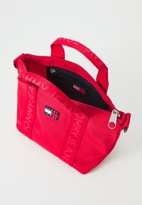 Czerwona torba Tommy Jeans na ramię z podwójnymi uchwytami, naszywką z logo z przodu oraz częściowo otwartym zamkiem błyskawicznym na górze, ukazującym wewnętrzną kieszeń z zamkiem.