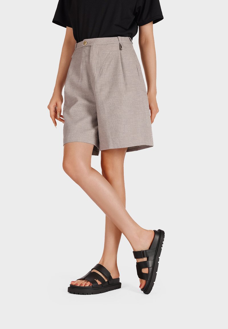 Pantaloni corti grigio chiaro fino al ginocchio con una tessitura strutturata e chiusura frontale con bottone, abbinati a sandali neri con lacci e suola spessa.