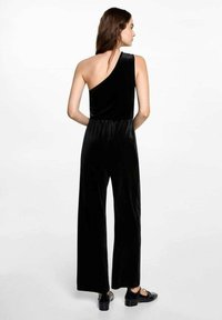 Black velvet jumpsuit with an asymmetrical one-shoulder design, wide-leg cut, and waistband detail. Flat shoes complete the look.