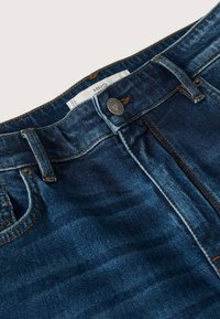 Tmavě modré džíny z denimu se zapínáním na knoflík, designem s pěti kapsami a kontrastním oranžovým prošíváním. Hladká textura, středně vysoký pas.