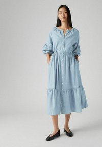 Vaaleansininen chambray-mekko, jossa on napit edessä, elastinen vyötärö ja kerrostettu hameosa. Hameessa on puffihihat ja sivutaskut. Mustat tasapohjaiset kengät.