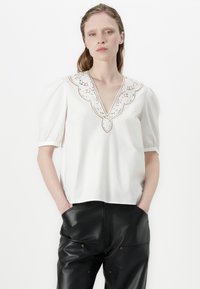 Blouse en coton blanc avec des manches bouffantes et un décolleté en V profond, présentant des détails en dentelle délicats autour de l'encolure. Texture lisse.