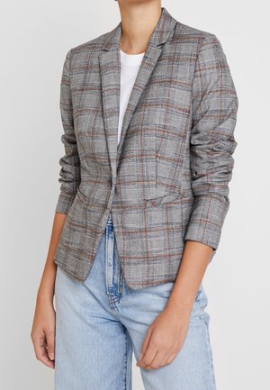Person trägt einen grau-braunen karierten Blazer mit gerafften Ärmeln über einem weißen Hemd und hellblauen Jeans.
