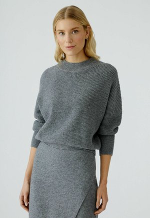 Blonde vrouw in een grijze geribde trui en bijpassende grijze kokerrok, staande tegen een effen lichte achtergrond.