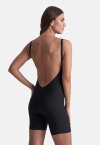 Mujer con un bodysuit ajustado negro sin espalda y con tirantes finos, de pie con una mano en la cadera, mirando a la derecha.