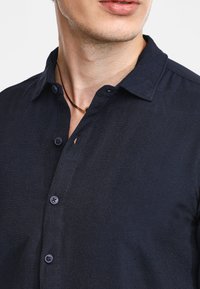 Camicia button-up blu scuro con colletto, tessuto testurizzato e bottoni frontali. Una collana marrone è visibile nella zona del collo.