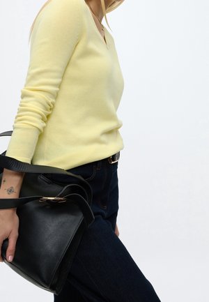 Femme portant un pull jaune et un jean foncé, tenant un sac à main en cuir noir sur son bras devant un fond uni.