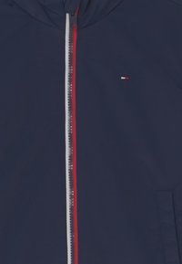 Granatowa kurtka przeciwdeszczowa z pełnym zamkiem błyskawicznym, z czerwonymi i białymi akcentami. Posiada małe logo oraz boczne kieszenie. Gładki, lekki materiał.