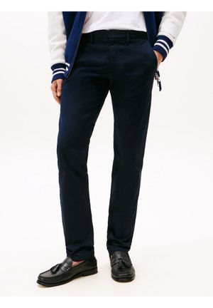 Uomo che indossa pantaloni slim fit blu navy, camicia bianca, giacca blu e bianca e mocassini neri con nappine, in piedi con una mano in tasca.