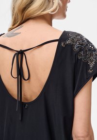 Blondynka z tatuażem ptaka, ubrana w czarną sukienkę z cekinowym, kwiatowym wzorem na ramieniu i wiązanymi ramiączkami na głębokim dekolcie na plecach.