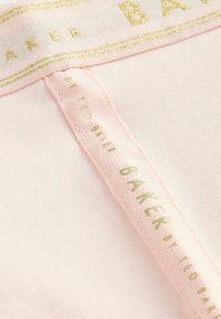 Svijetlo ružičasta tkanina s glatkom teksturom ima oznaku s zlatnim akcentom na kojoj piše "BAKER" i "by TED BAKER" u komplementarnoj zelenoj boji.