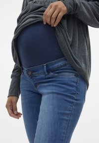 Grå långärmad överdel draperad över en blå denim graviditetsband, kombinerad med tajta ljusblå jeans med knappstängning och fickor.