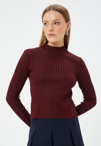 Burgunderfarbener, gerippter Pullover mit langen Ärmeln und hohem Hals. Er zeichnet sich durch sein tailliertes Design und den strukturierten Stoff aus, der mit einem dunklen Rock kombiniert wird.