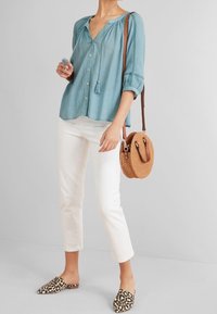 Chemise bleu clair à boutons avec manches trois-quarts et détail à pompons. Pantalon blanc court, chaussures à enfiler imprimé léopard, et sac rond en paille.