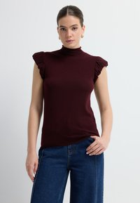 Top sin mangas burdeos con cuello alto y detalles de hombros con volantes, hecho de una tela suave, combinado con jeans de mezclilla azul de pierna ancha.