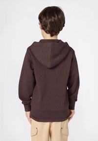 Barn iført en mørkebrun hettegenser vendt bort fra kameraet og beige cargo-bukser, stående mot en enkel hvit bakgrunn.
