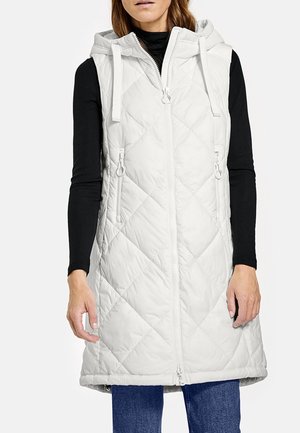 Frau trägt ärmellose weiße gesteppte Kapuzenweste über schwarzem Langarmshirt und blauer Jeans, steht vor weißem Hintergrund.