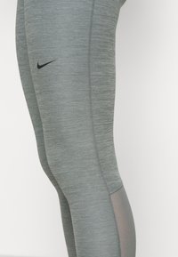 Szare legginsy sportowe z teksturowanym wykończeniem, z czarnym logo Nike, siateczkowym panelem po dolnej stronie oraz kontrastowym przeszyciem.