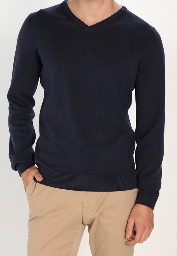 Homme portant un pull bleu marine à col en V et manches longues et un pantalon beige, une main dans la poche, sur un fond uni.
