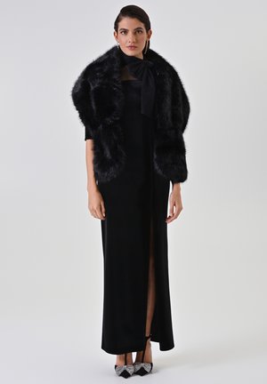 Abito in velluto nero con uno spacco fino alla coscia, abbinato a un stole in pelliccia sintetica nera e tacchi neri decorati con dettagli a fiocco.