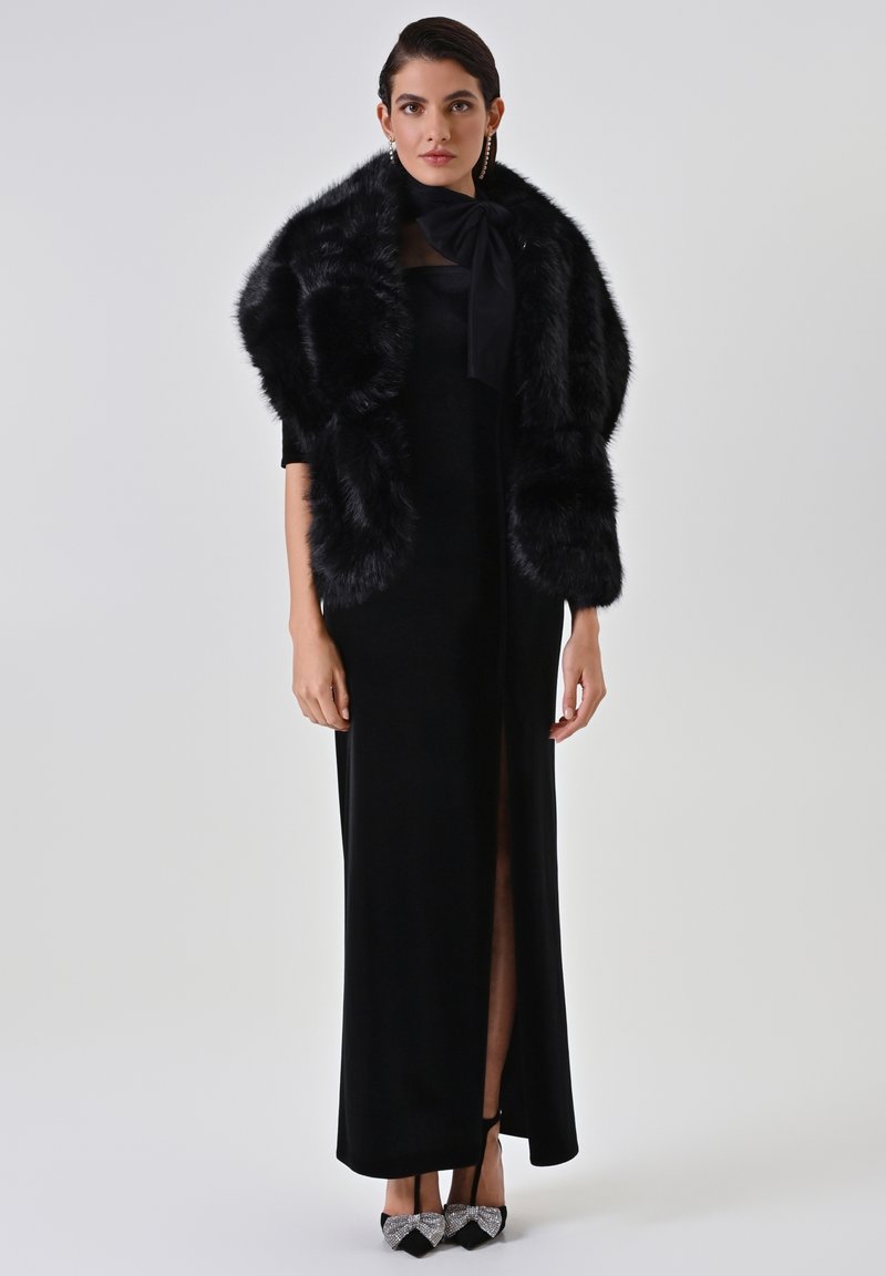 Abito in velluto nero con uno spacco fino alla coscia, abbinato a un stole in pelliccia sintetica nera e tacchi neri decorati con dettagli a fiocco.