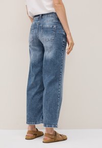Une personne se tient de dos face au spectateur, portant un pantalon en jean large bleu et des sandales marron, sur un fond neutre.