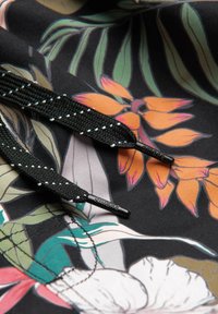 Des lacets noirs avec des coutures blanches reposent sur un tissu floral coloré présentant des feuilles et des pétales orange, blancs, verts et roses.