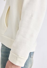 Kremfarget genser med hette laget av myk stoff, med en frontlomme, ribbede mansjetter og et lite hull på ermet. Kombinert med jeans.