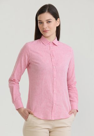 Nő hosszú sötét hajjal, aki rózsaszín, gombos, hosszú ujjú inget és bézs nadrágot visel, zsebeiben tartja a kezét, semleges háttér előtt áll.