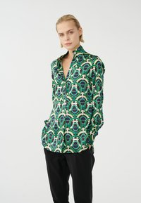 Grüne gemusterte Bluse mit geometrischen Kreisen, Knopfverschluss vorne, langen Ärmeln und lockerem Sitz, kombiniert mit schwarzen Hosen.