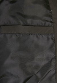 Black nylon jacket fabric displaying a smooth texture, with a rectangular pocket on the left side and visible stitching details.