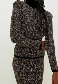 Donna che indossa un vestito a maglia con stampa leopardata marrone e nera, maniche lunghe e rifiniture nere ai polsini e in vita, in piedi contro uno sfondo neutro.