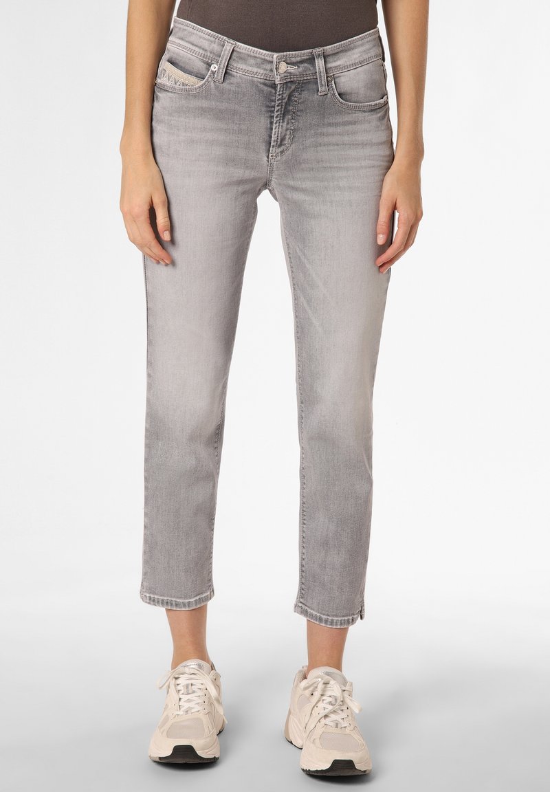 Femme portant un jean court gris clair et des baskets beiges, debout devant un fond blanc uni.