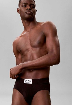 Athletic man wearing black Calvin Klein briefs, standing against a plain gray background, looking to the side with arm bent.