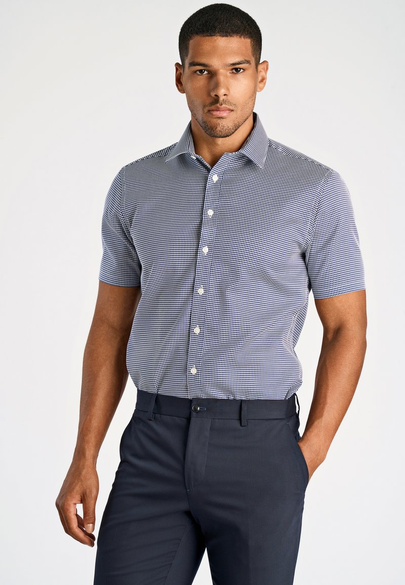 Camicia a maniche corte con bottone, a fantasia quadrata in navy e bianco, abbinata a pantaloni slim-fit navy. Texture liscia, design su misura.