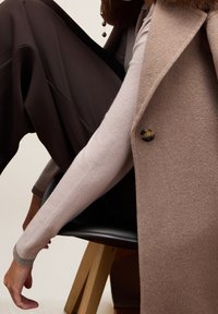 Beżowa kamizelka z mieszanki wełny z brązowym guzikiem, nałożona na jasnoróżowy dopasowany top. Widoczne ciemnobrązowe szerokie spodnie. Gładkie faktury.